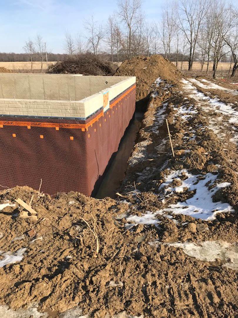 Foundation trench surrounded by piles of dirt and patches of snow, bordered by trees at a residential construction site