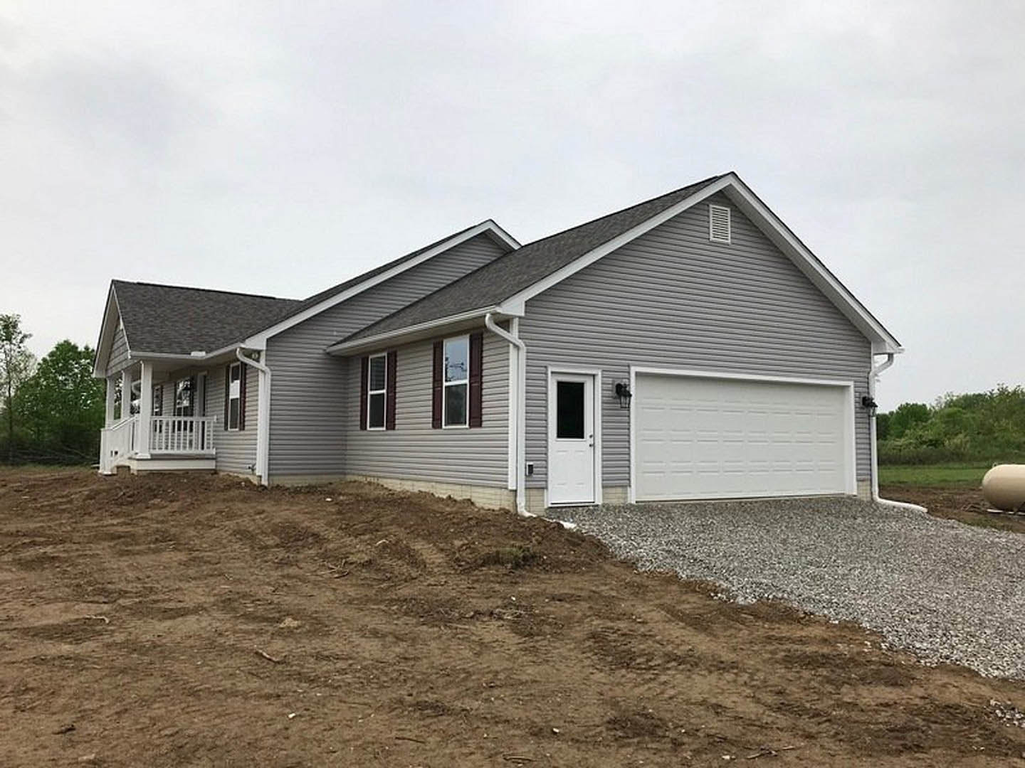 Two-story home with light-colored siding, attached garage with white paneled door and window, gravel driveway, dirt hill to the side, man standing near garage, scattered trees in