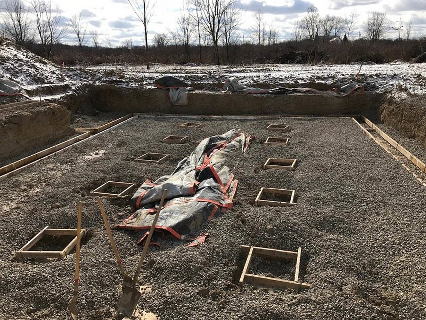 Gravel-covered construction site with wooden square frames and tarp, surrounded by leafless trees under cloudy winter sky