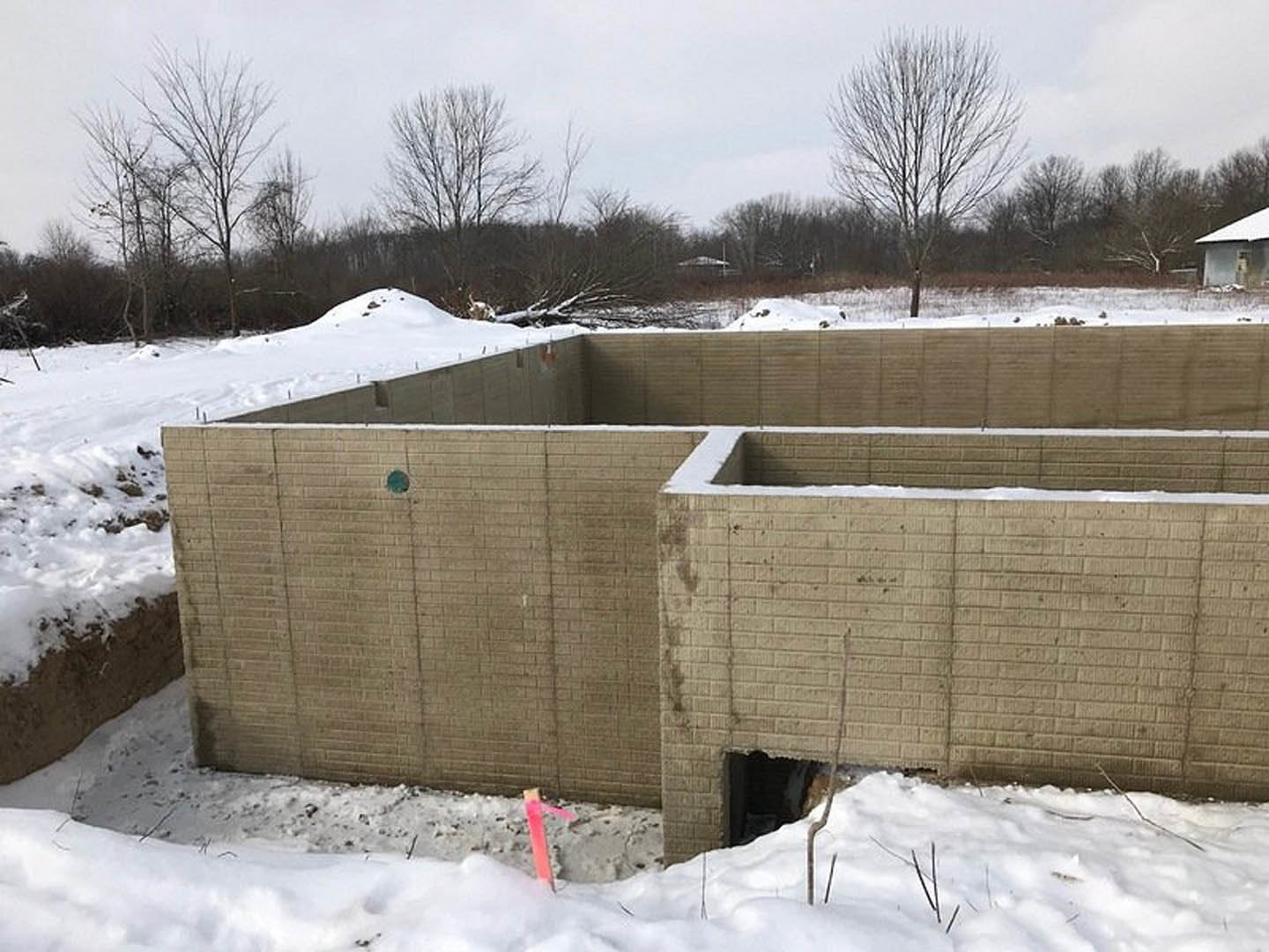 Concrete foundation and walls surrounded by snow, leafless trees in the background, pink construction tape marking the site, winter sky overhead.