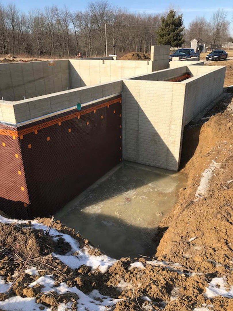 Concrete foundation with exposed hole in the ground, brown wall surface, leafy tree, concrete wall featuring a hole, pile of dirt and snow, power lines overhead, winter landscape