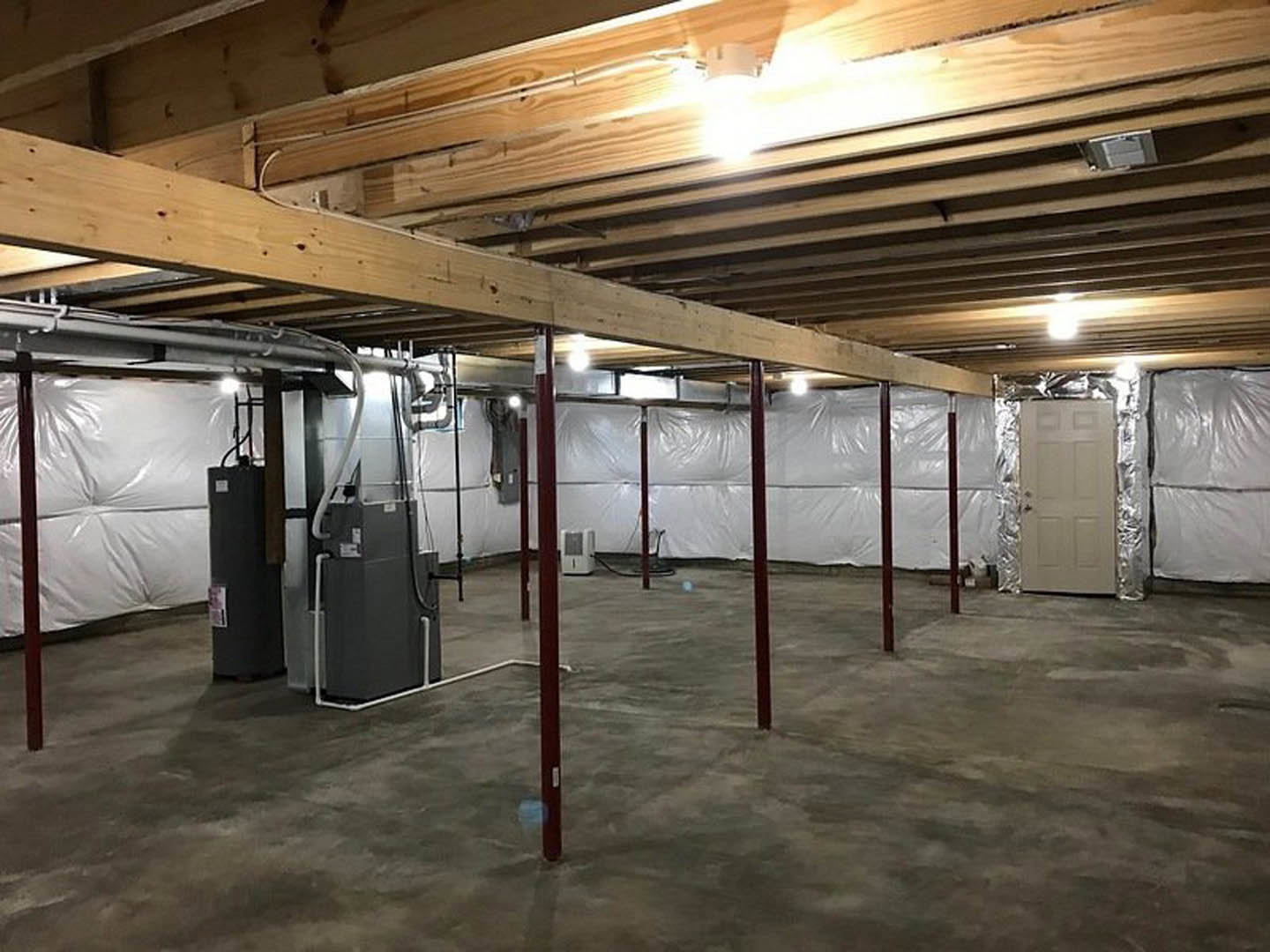 Basement room with white painted walls, exposed wooden ceiling beams, steel support post, white door with metal doorknob, visible mechanical equipment and pipes, concrete floor