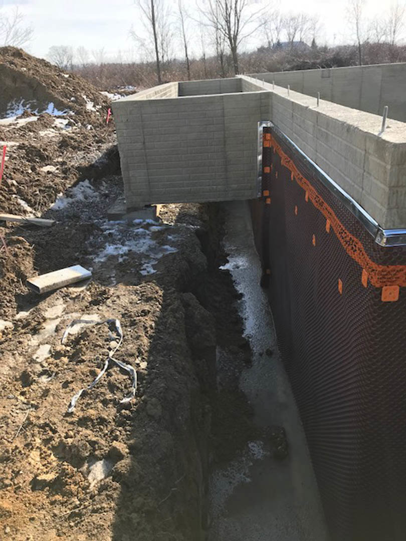 Concrete foundation with exposed hole in the ground, black mesh covering along wall, white construction object in foreground, outdoor setting with trees and sky visible
