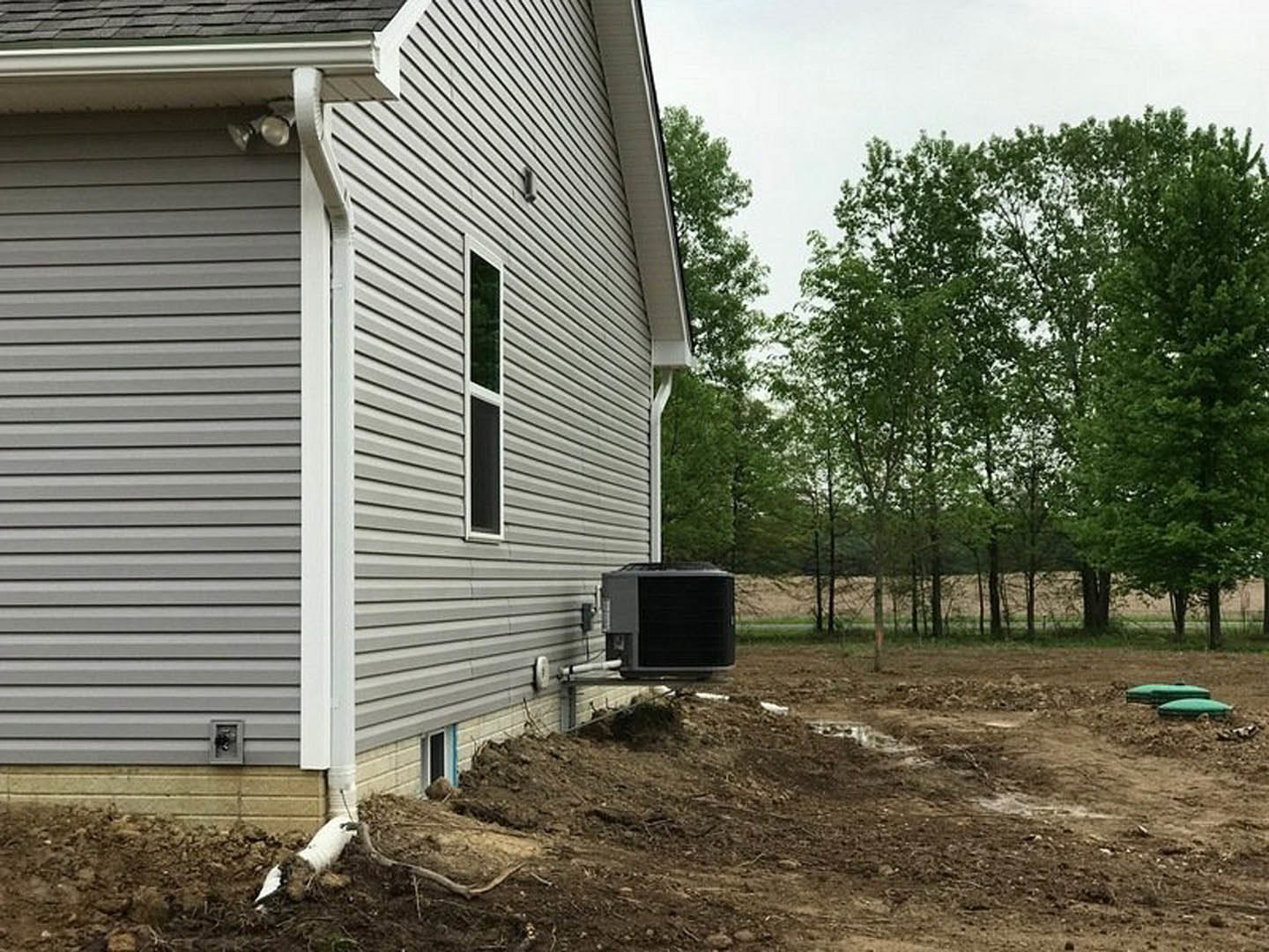 Side view of a custom home with light-colored siding, large air conditioning unit mounted outside, square windows, and mature trees in the yard.
