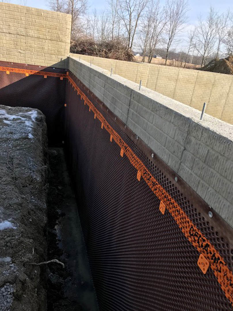 Concrete exterior wall with circular hole, surrounded by black mesh, adjacent to pile of rocks and winter trees under cloudy sky.