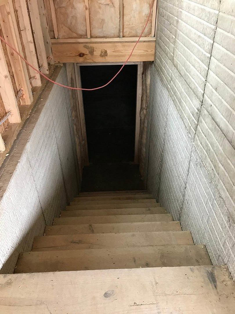 Wood plank staircase with black metal railing descending to concrete basement floor, surrounded by white walls and minimal decor.
