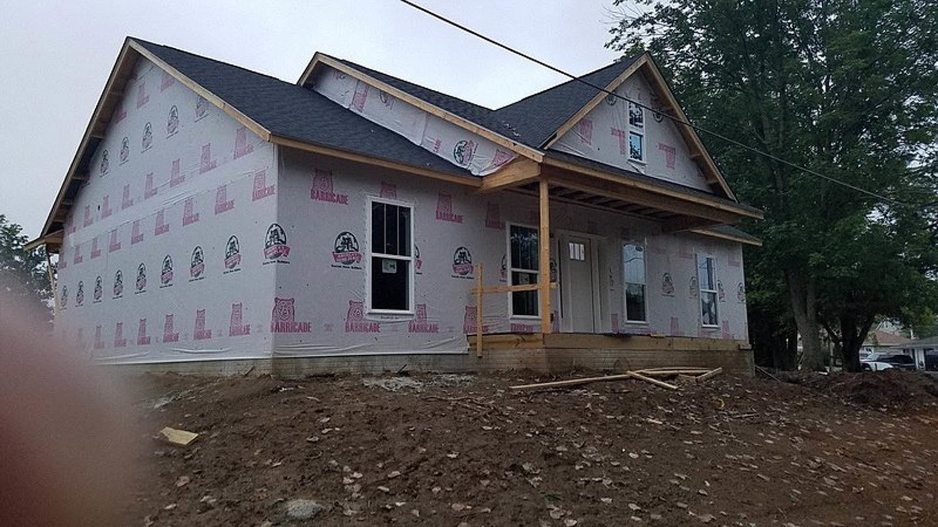 Partially built house with exposed plywood walls, red tape sealing seams, white-framed window, and roof covered in protective plastic sheeting