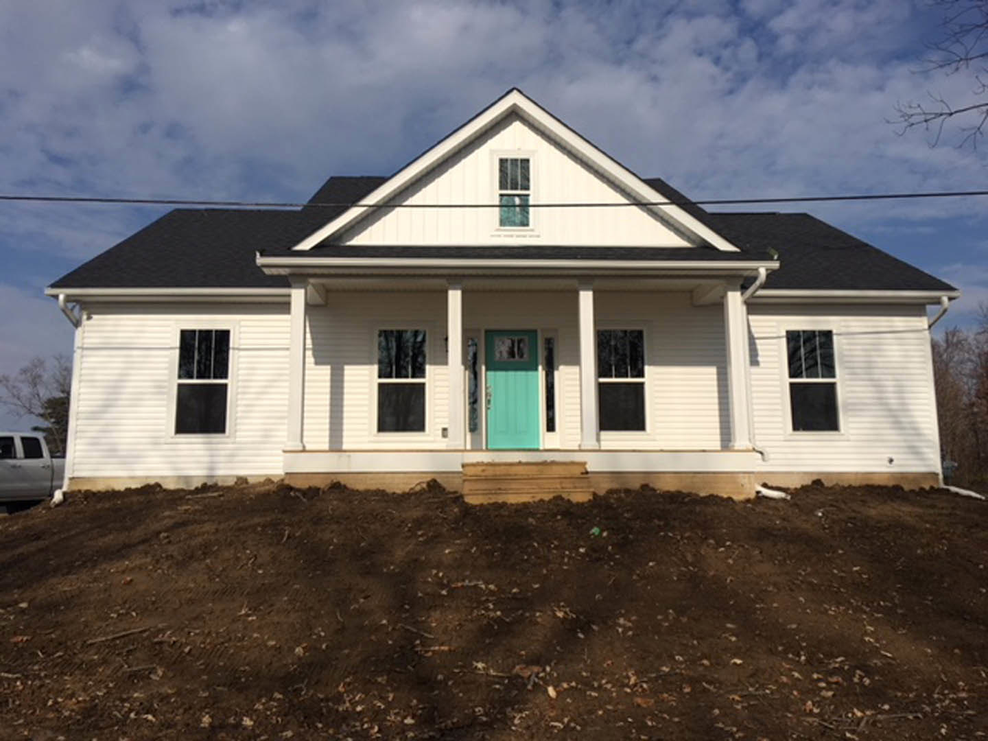 White cottage-style home with blue front door, white siding, row of windows, dirt and leaves on ground, cloudy sky overhead