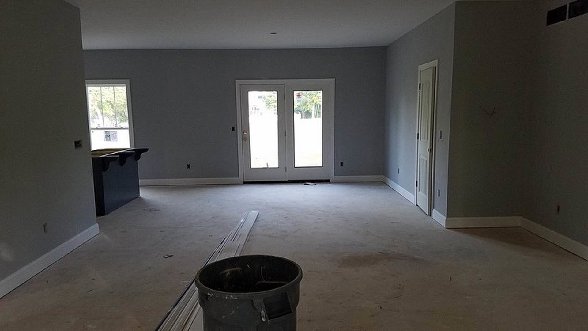 White-walled room with light wood flooring, a black paint bucket on the floor near a window, unfinished plaster surfaces, and a closed door.