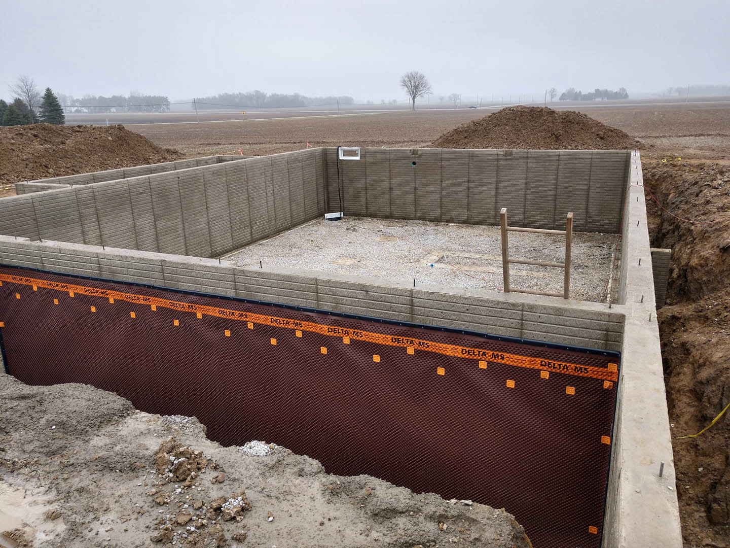 Gravel construction site with fenced excavation, wooden ladder, exposed wall, tree in grassy field, blue sky with clouds