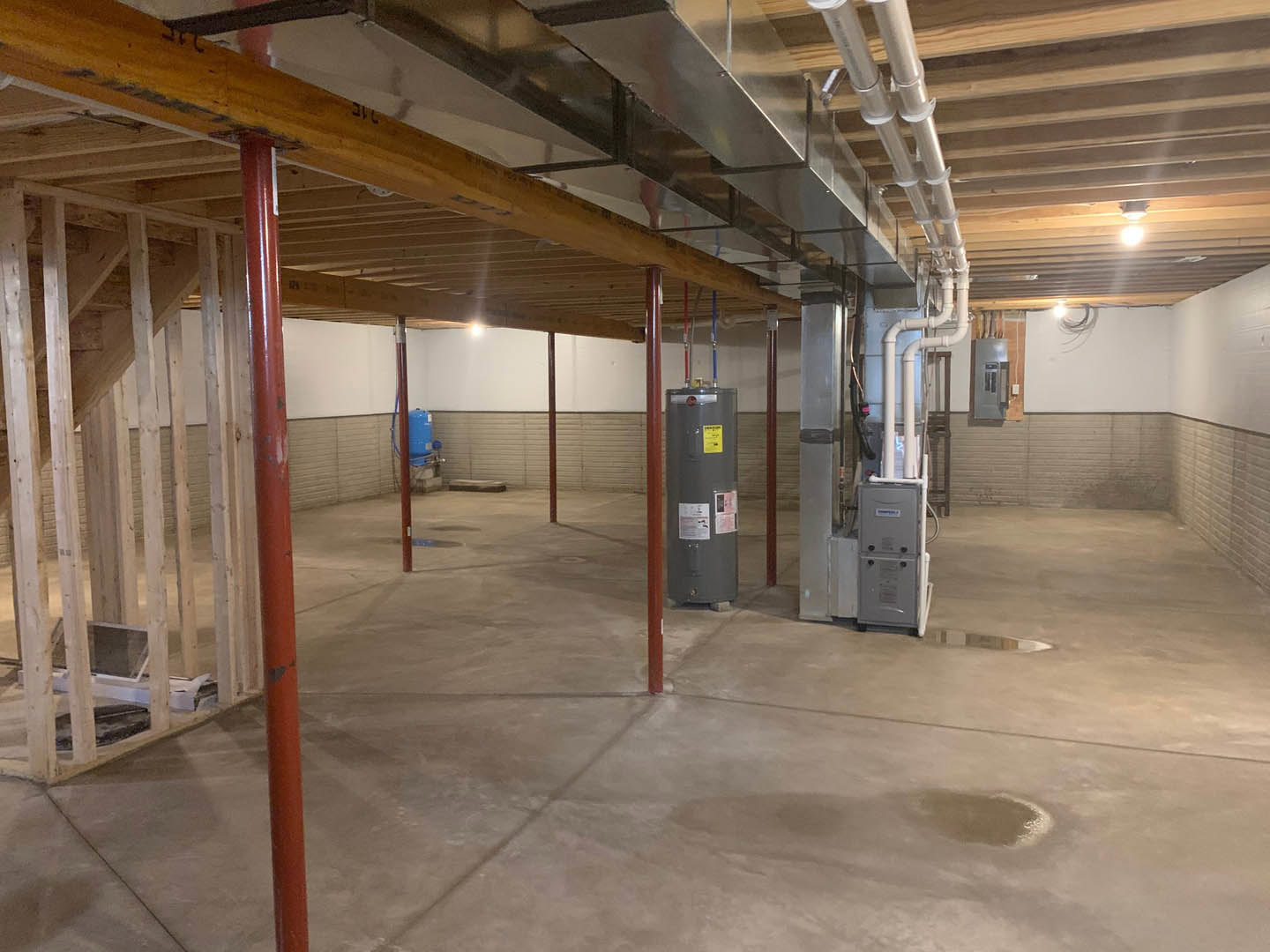 Exposed pipes and cylinders with yellow labels, concrete floor with red poles, wooden ceiling beam, grey walls, and construction materials in an unfinished room