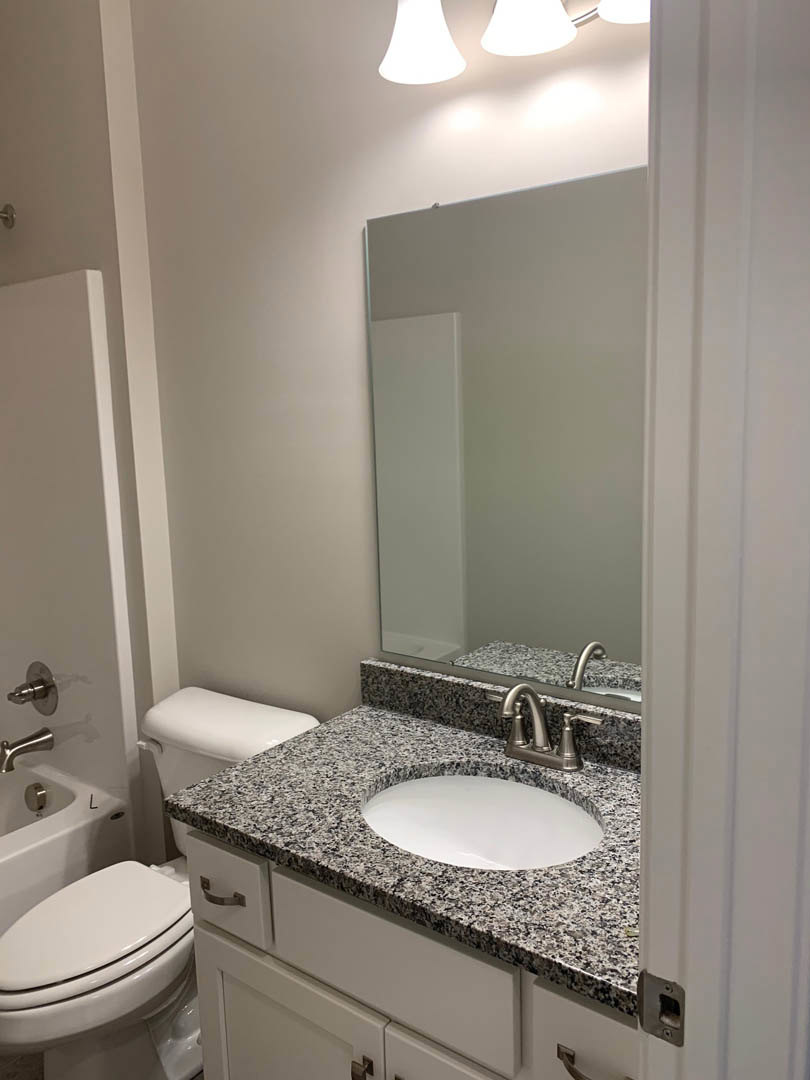 Marble countertop with oval sink, chrome faucet, white cabinet, wall-mounted mirror, and toilet against white tile walls