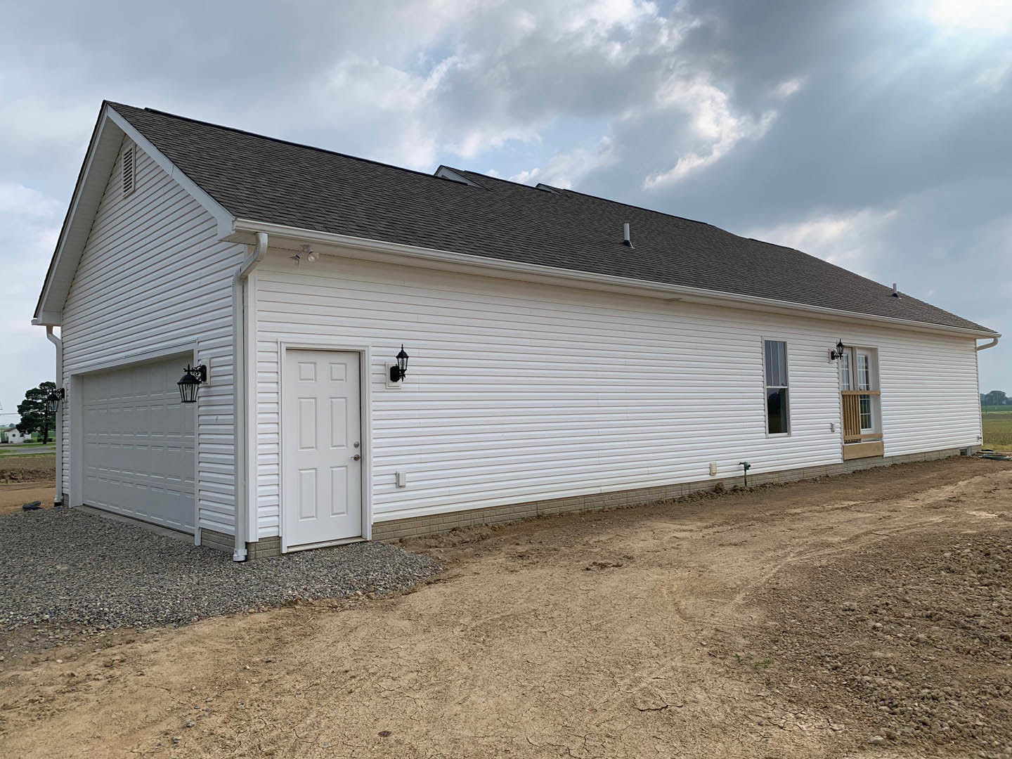 White house with a gabled roof, white door featuring a silver handle, adjacent dirt and gravel path, window with wooden slat, exterior wall-mounted light, cloudy sky overhead.