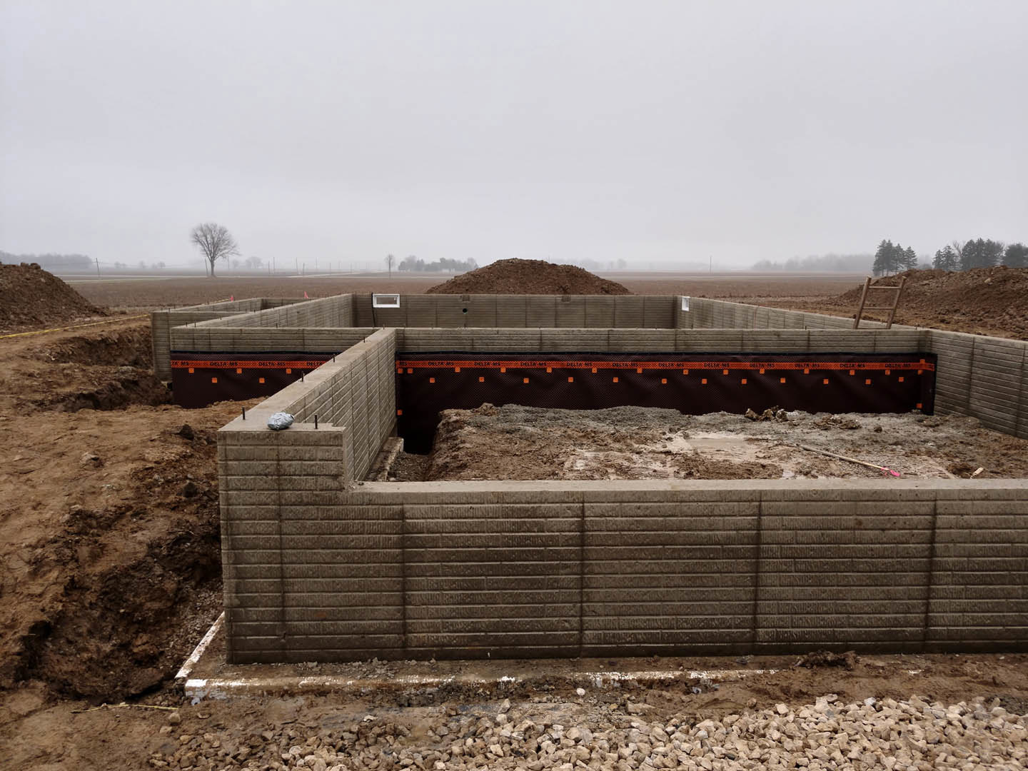 Concrete foundation with exposed rebar, surrounding dirt and rocks, partially built wall with opening, overcast sky, tree in background