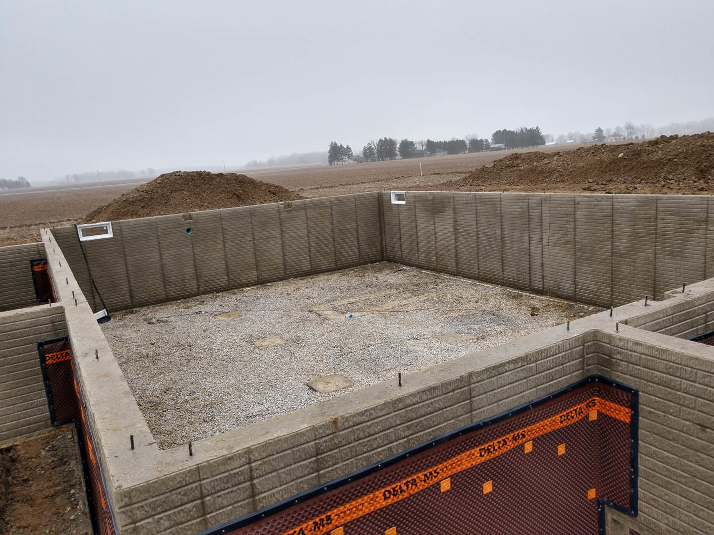 Concrete foundation with exposed gravel and dirt, unfinished wall section, window opening, distant field and trees, construction site signage