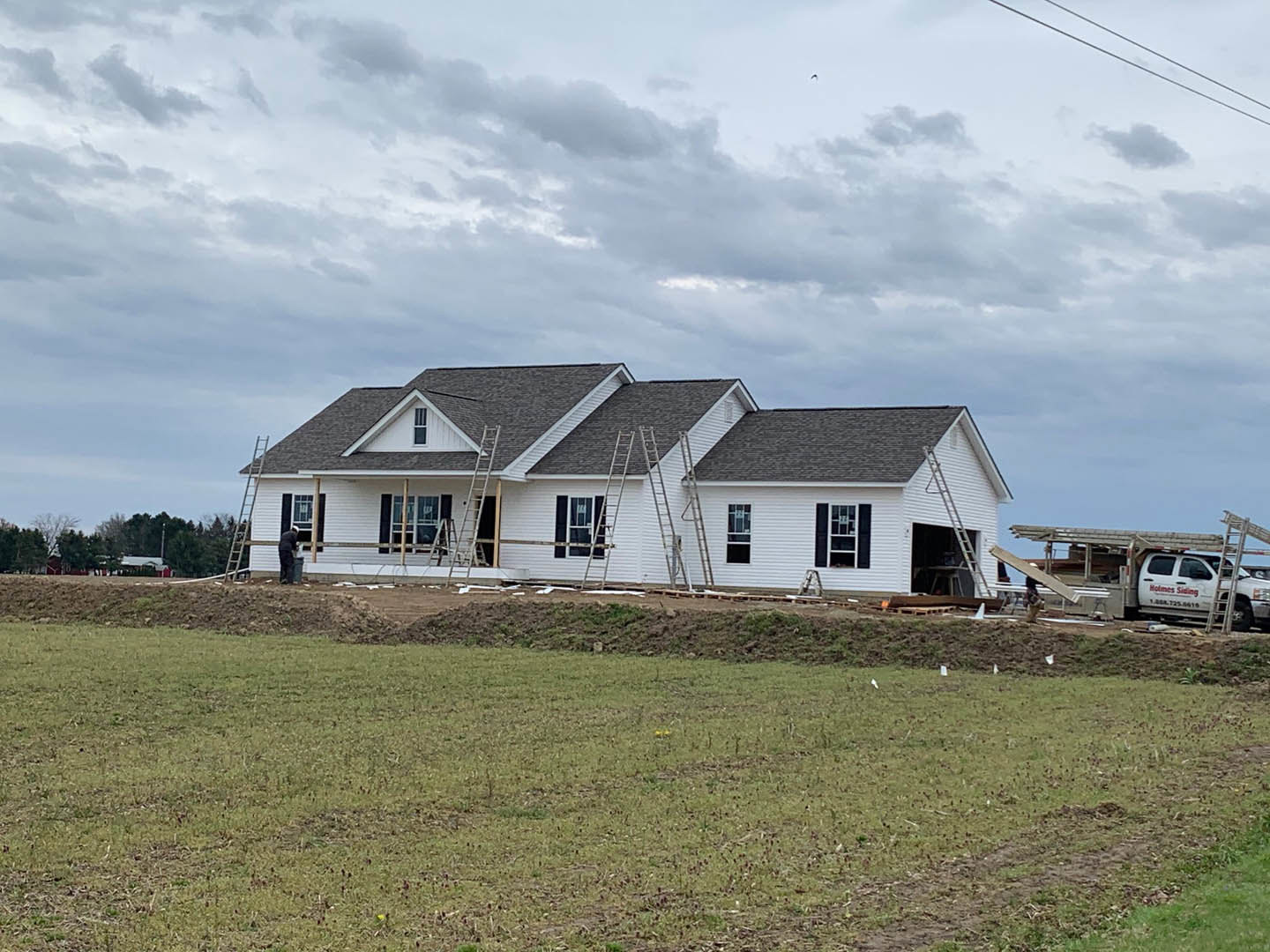 Wood-framed house under construction with ladders propped against the roof, grassy lot in foreground, Southfork Ranch visible in the background, white truck parked beside