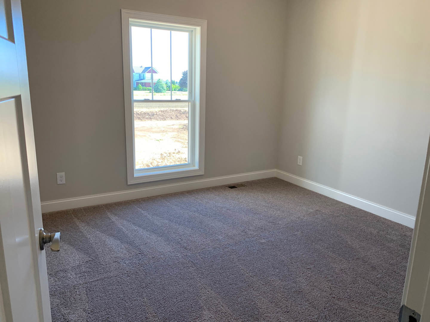 Neutral-toned carpeted room with white walls, large window overlooking a field and neighboring house, white door with chrome handle, and ceiling with recessed lighting
