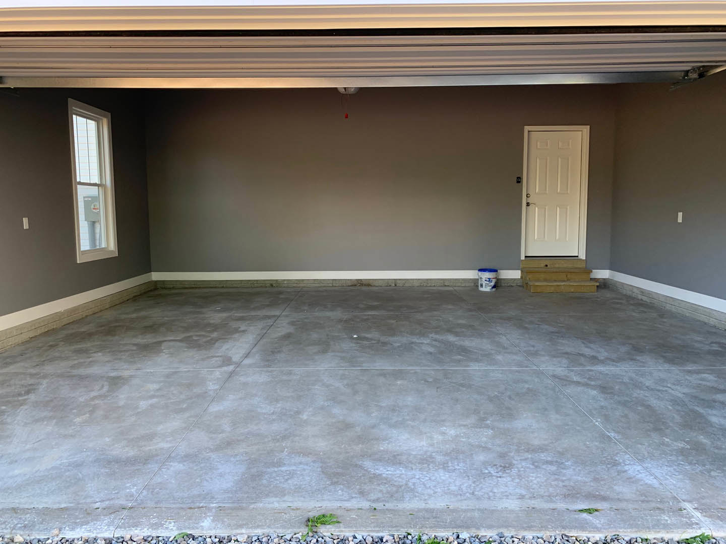 Garage interior with concrete floor, white entry door with silver handle, paint bucket, white container with blue lid, window with white frame, and close-up of wooden staircase