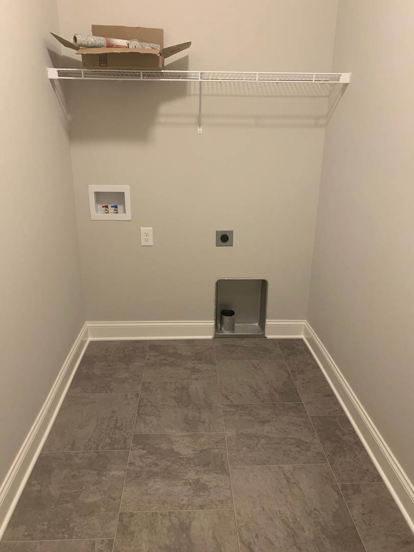 Small closet with brown tile flooring, white trim, metal shelving, cardboard box with a roll of paper, white wall featuring a white shelf and red and blue faucet, metal can placed