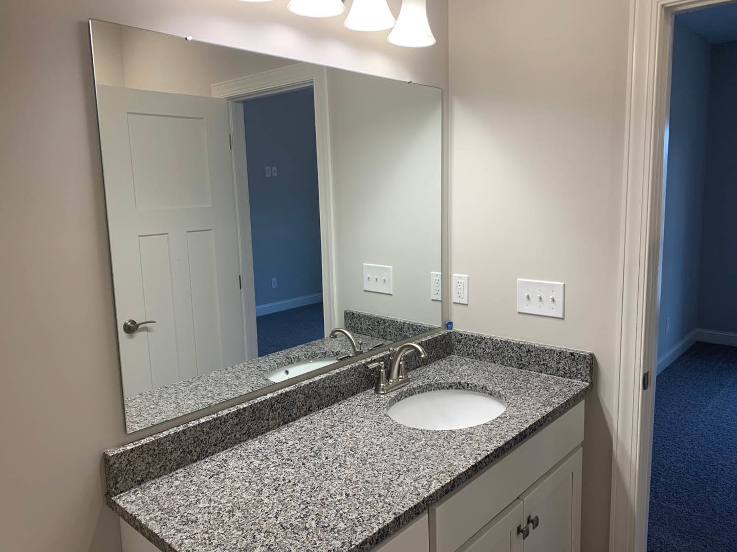 Bathroom with stone countertop featuring two white sinks, large wall mirror, white wall switch with three knobs, and close-up of brushed metal door handle.