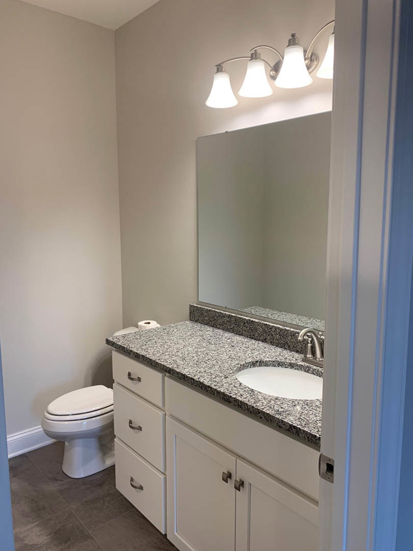 Marble countertop with undermount white sink, chrome faucet, white toilet, white cabinetry, wall-mounted mirror, and light fixture in a tiled bathroom.