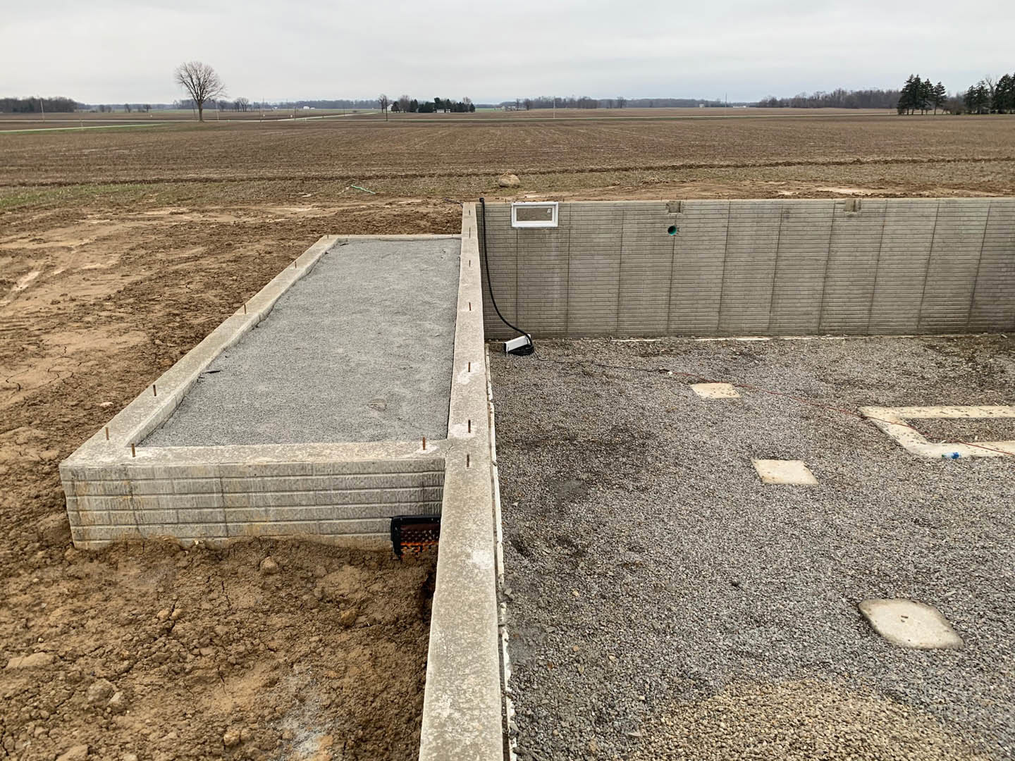 Concrete foundation with grey concrete wall, blue pipe, gravel ground, leafless tree, and hole in the wall; outdoor construction site with soil and grass.