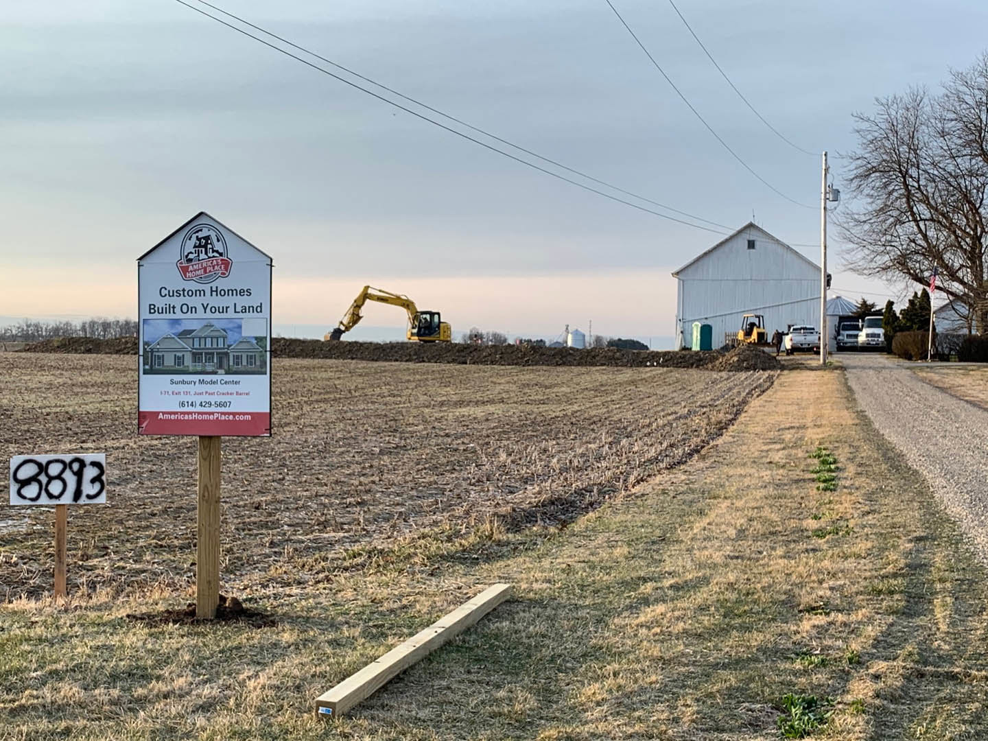 White sign with black numbers mounted on a pole in grassy field, construction site in background featuring white building, yellow and black tractor, white truck, and scattered