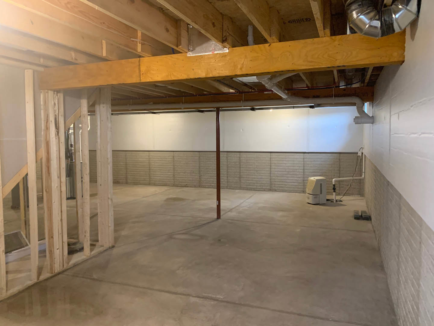 Open room with exposed wooden ceiling beam, concrete floor, white vented rectangular object, visible metal pole, and white pipe with attached black pipes along wall.