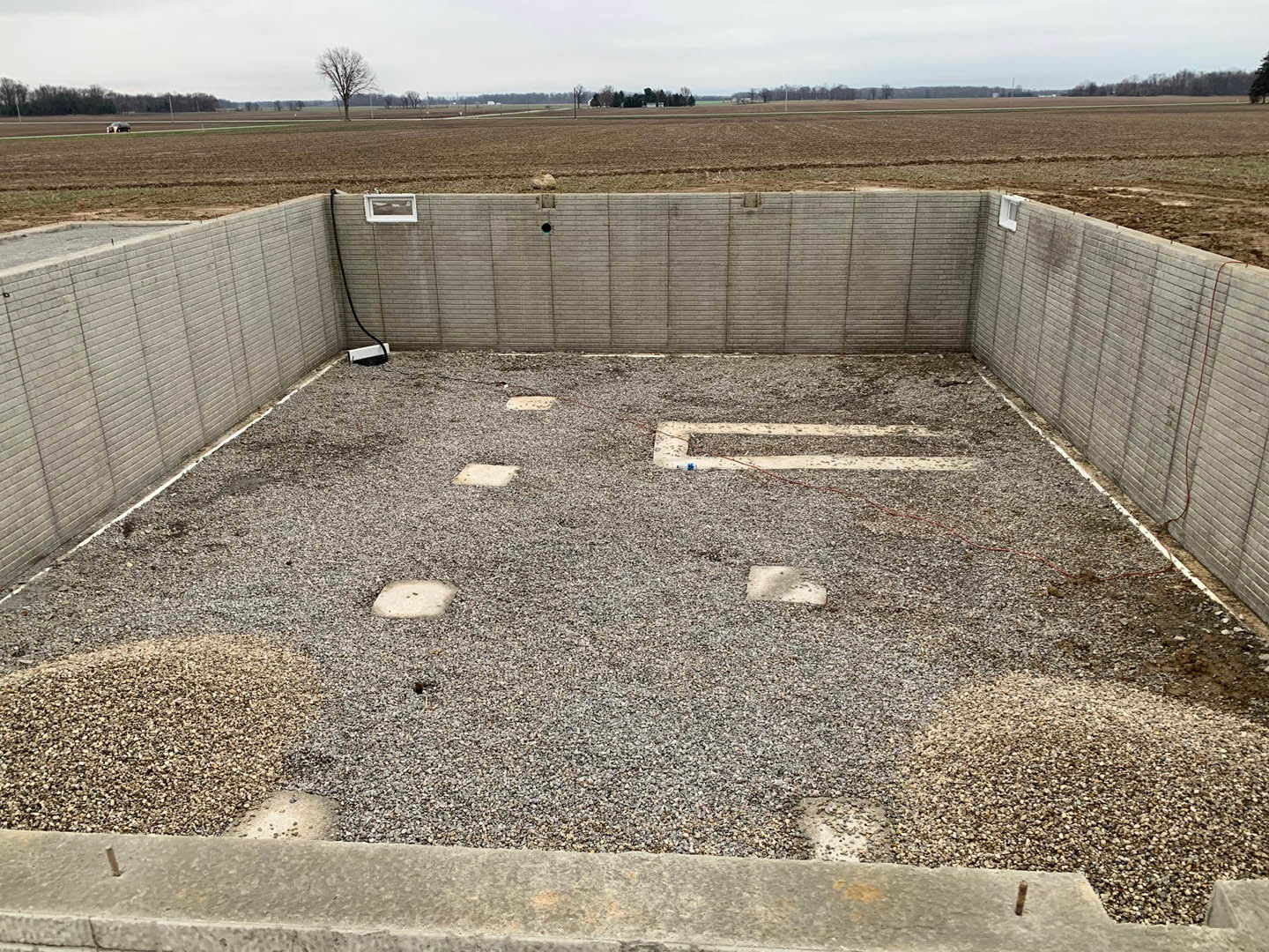 Concrete foundation surrounded by gravel, expansive grassy field in background, leafless tree, grey brick wall, and scattered stones visible.