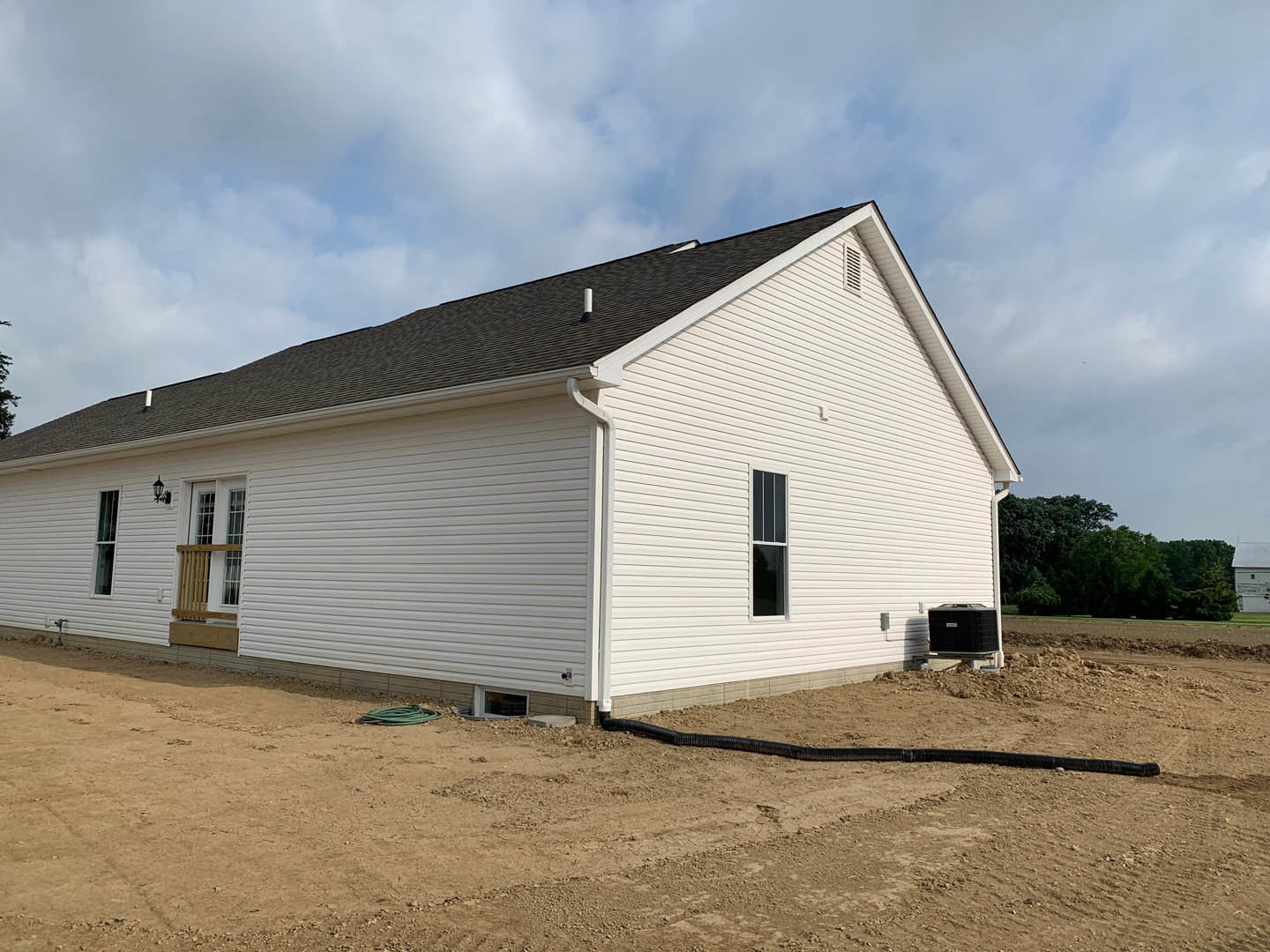 White house with black roof, white-framed windows, wooden railing, dirt yard, distant trees under cloudy sky