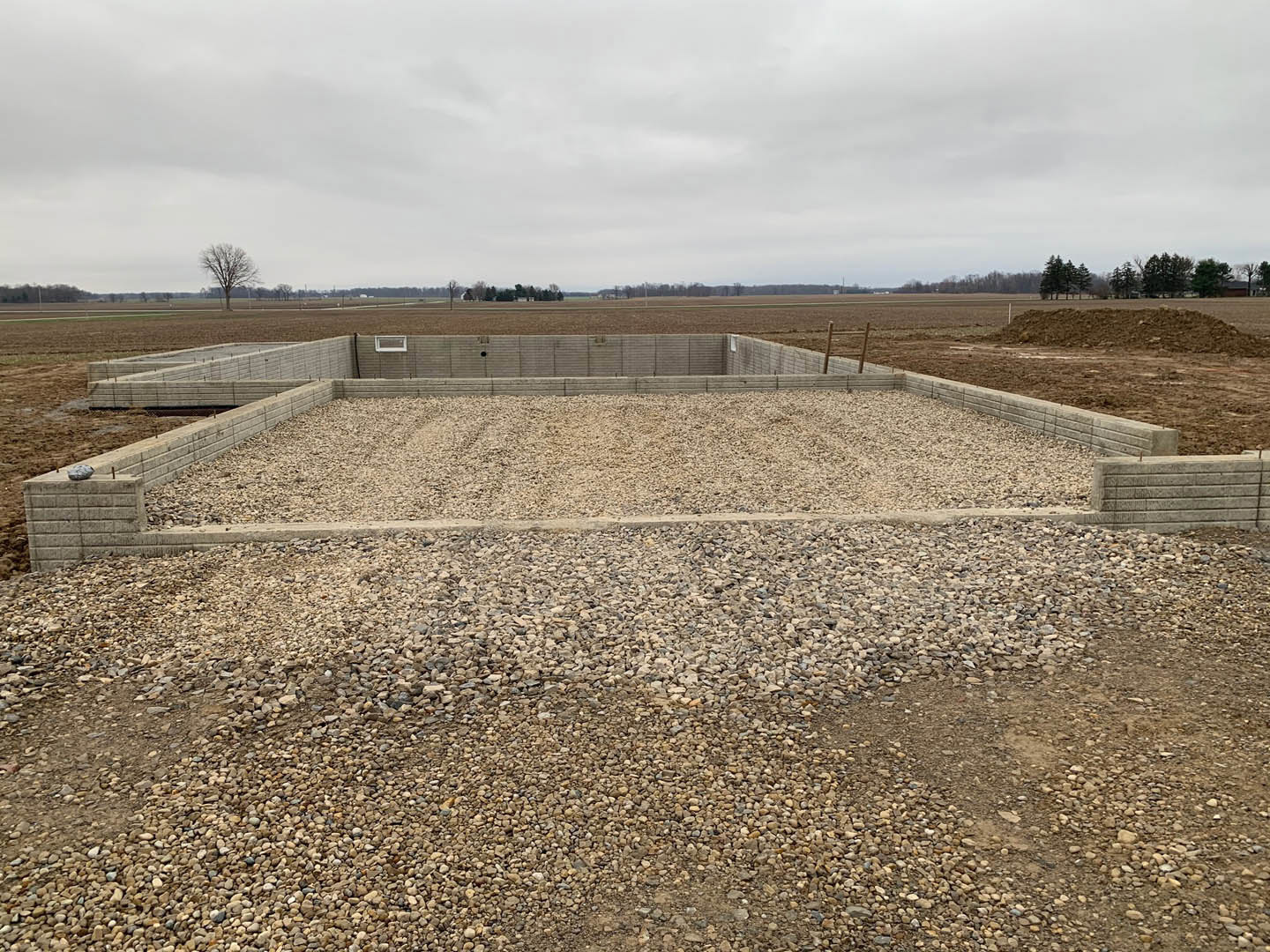 Gravel and concrete foundation slab surrounded by grass and soil, with scattered trees in the background under a cloudy sky