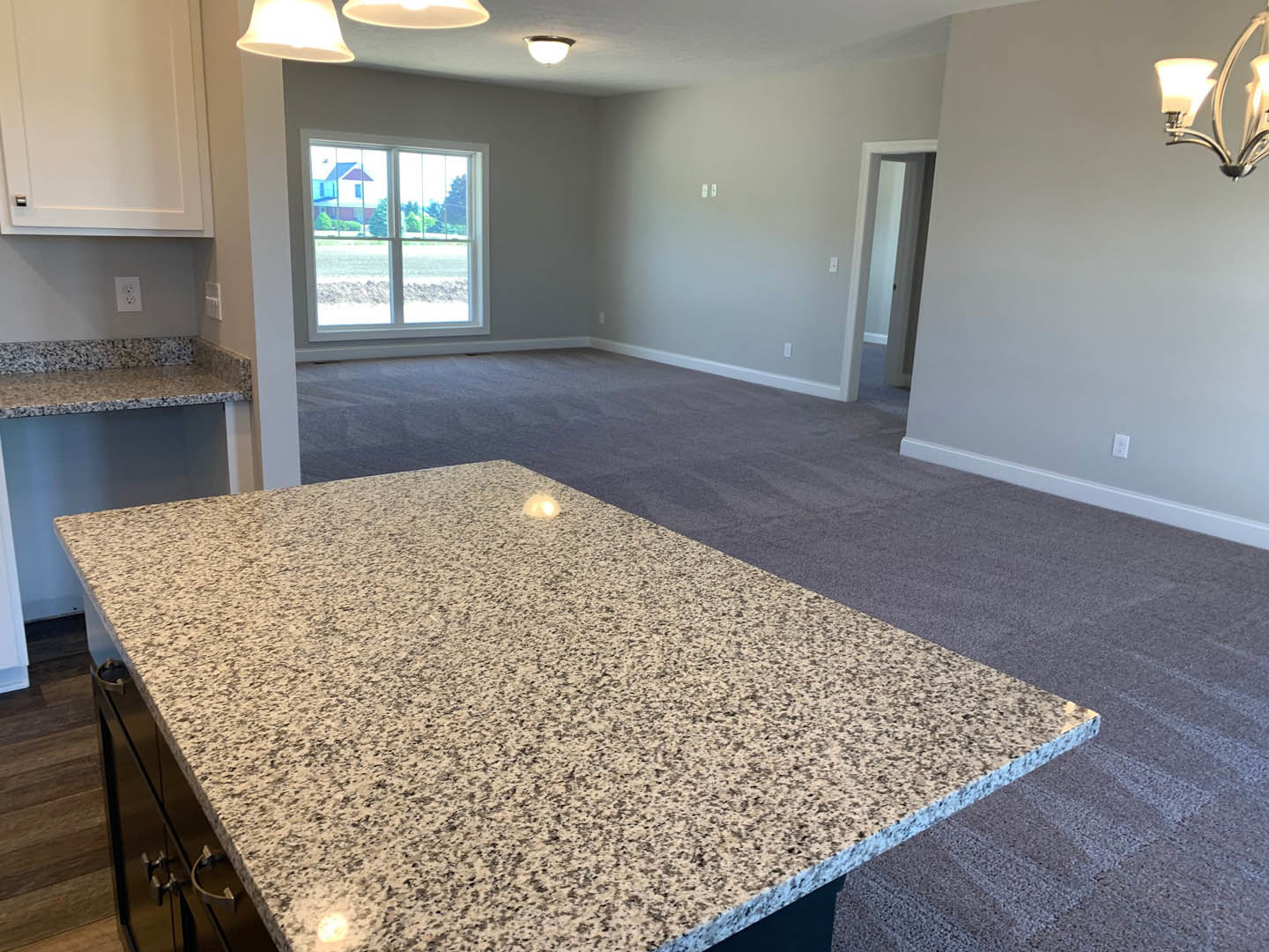 Open-concept kitchen and living room featuring granite countertops, tile flooring, white cabinetry, stainless steel sink, modern light fixtures, and a window overlooking the