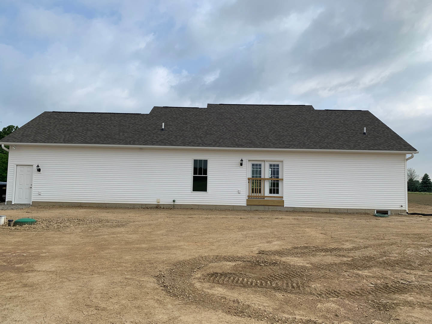 White house with black roof, tire tracks in dirt yard, balcony with railing, large windows, cloudy sky overhead