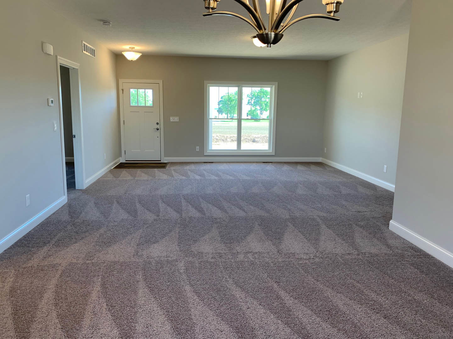 Carpeted room with white paneled door, window overlooking trees, decorative chandelier hanging from plaster ceiling, wall vent visible