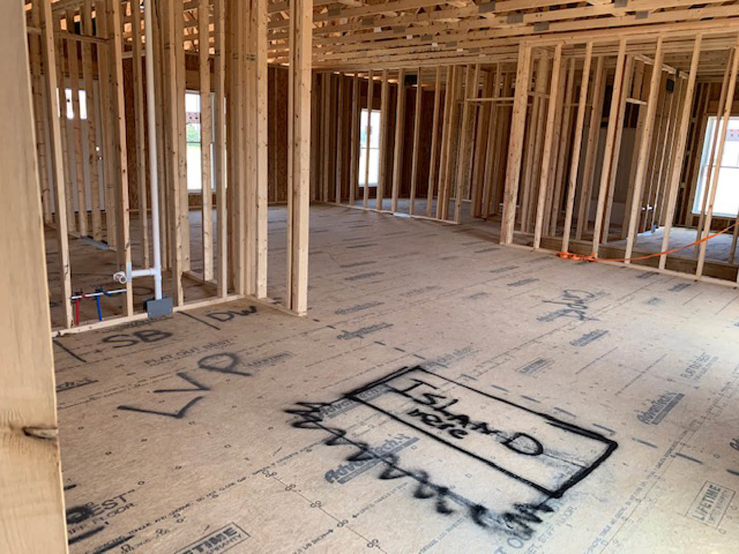unfinished room with exposed wood framing, plywood subfloor marked with handwritten notes, large window letting in natural light