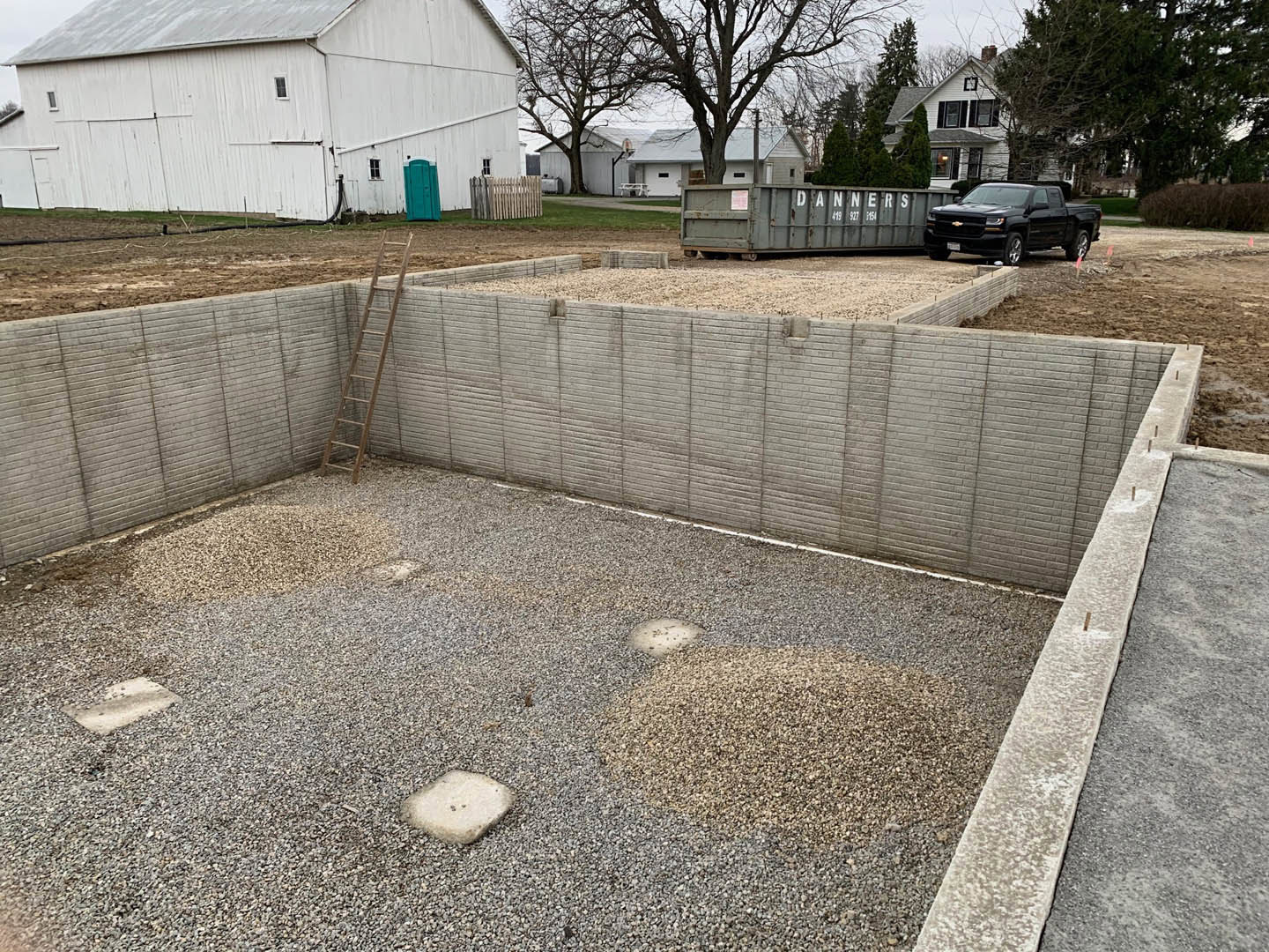 Modern custom home with concrete foundation, light-colored siding, black SUV parked in driveway, white barn with metal roof in background, gravel yard, and mature trees surrounding