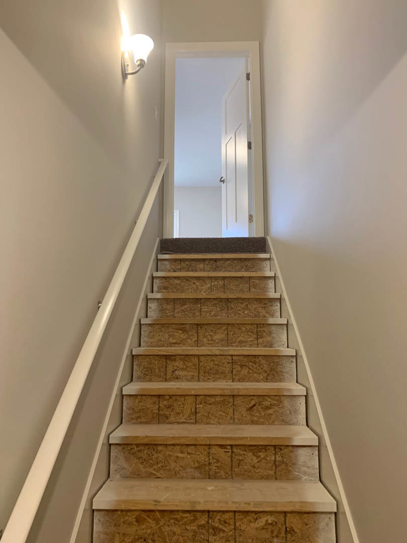Wood staircase with white plaster walls, silver handrail, ceiling light fixture illuminated, white door with silver handle, wooden shelf mounted on wall, light-colored flooring and