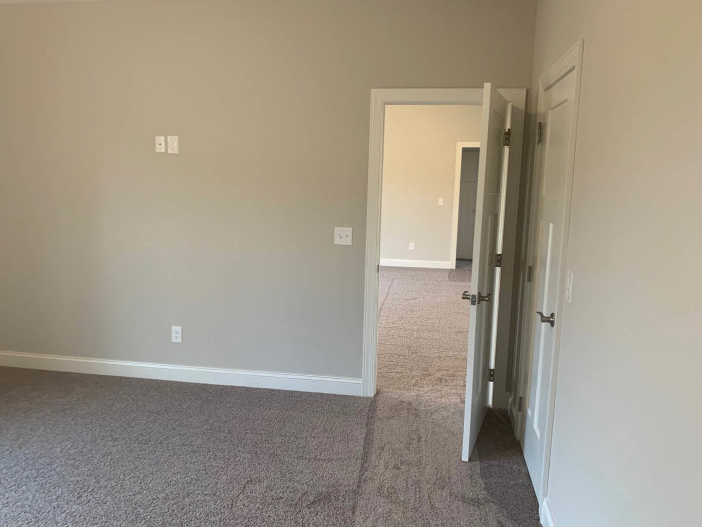 White painted door open against smooth white wall, beige carpet flooring, electrical outlet visible near baseboard, ceiling with recessed lighting.