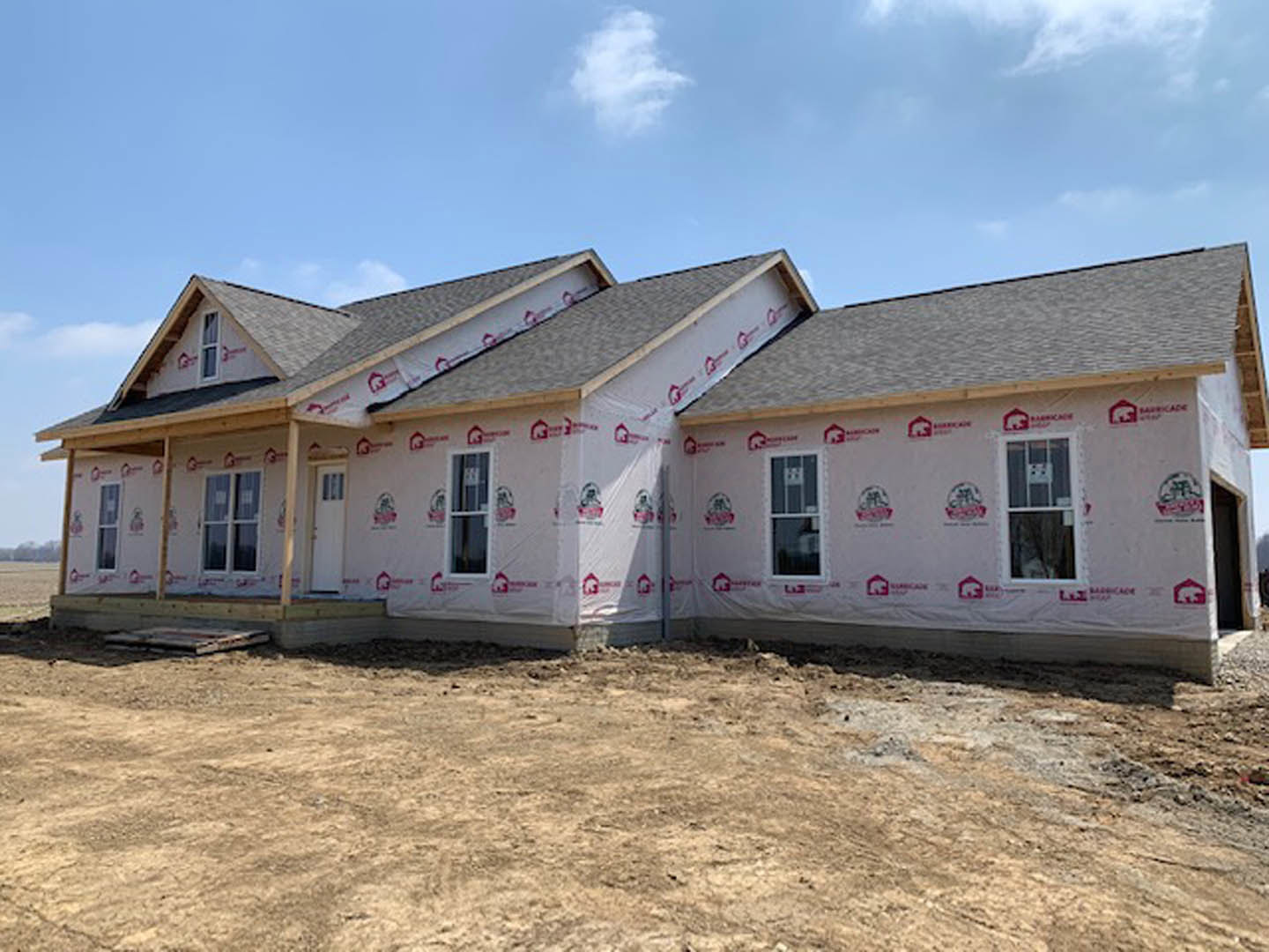 Partially built house with exposed framing, red tape sealing seams, plastic sheeting covering exterior walls, unfinished roof, and windows installed under cloudy sky