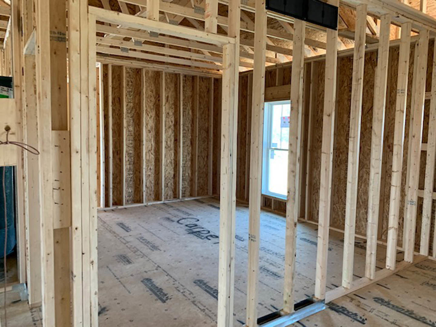 Wood-framed room under construction with exposed beams, white-framed window, black screen resting on plywood surface, and visible insulation materials