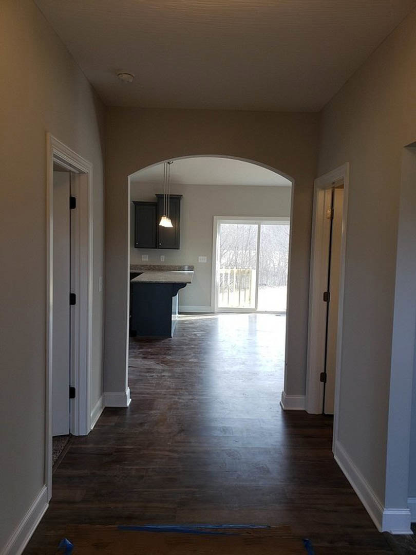 Hallway with dark wood flooring and white trim, leading to a kitchen and dining area; large window overlooks snowy forest; close-up details of a stone countertop, modern light