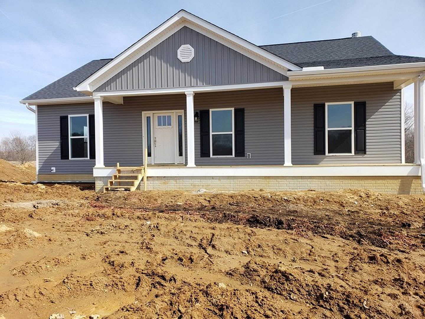 Gray-sided custom home under construction with white vent, white door with glass panes, black shuttered window, covered porch with stairs, and exposed dirt yard foreground