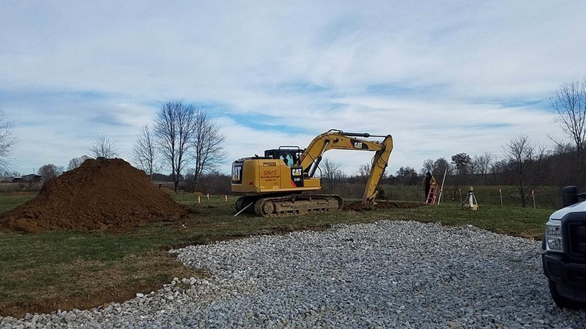 Yellow excavator with large wheels parked on soil in open field, cloudy sky overhead, trees in background, truck partially visible in foreground