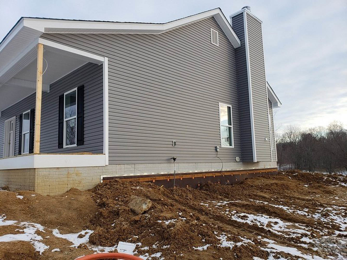 Two-story home under construction with white-framed windows, gray siding, exposed chimney, dirt and snow piles in the yard, garden hose on ground, and leafless trees in background.
