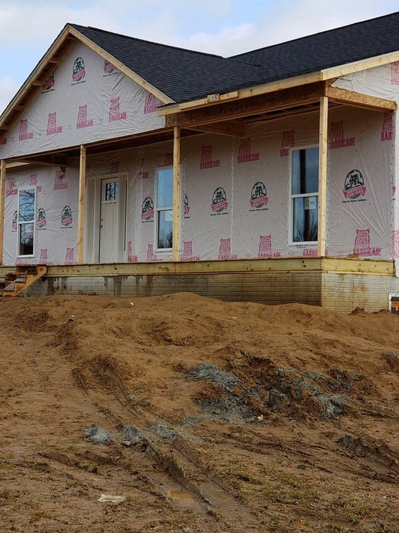 Partially built house with white siding, plastic sheeting covering windows, dirt mound and tire tracks in foreground, cloudy sky overhead
