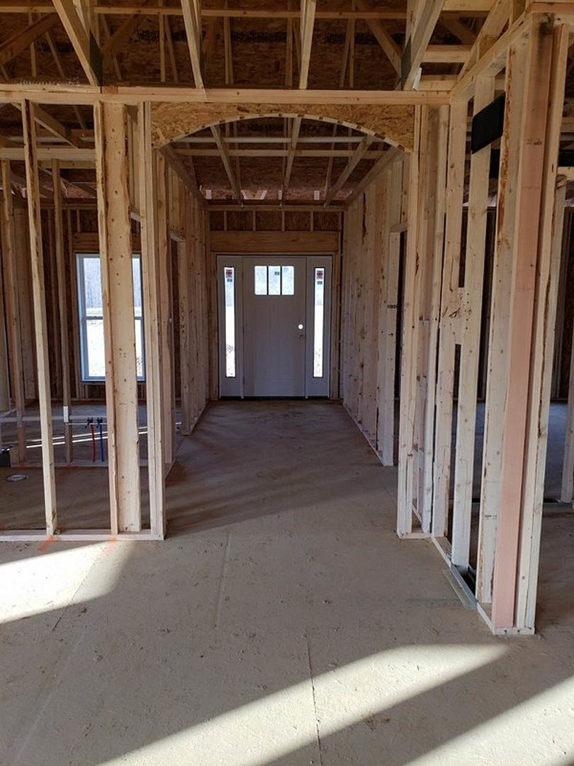 Framed hallway with exposed wooden beams, unfinished drywall, and a newly installed door; row of windows with construction signage visible.