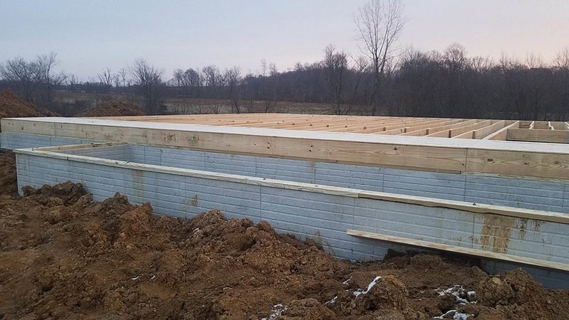 Partially built house with exposed wooden beams, foundation surrounded by soil, leafless tree and cluster of trees in background under open sky