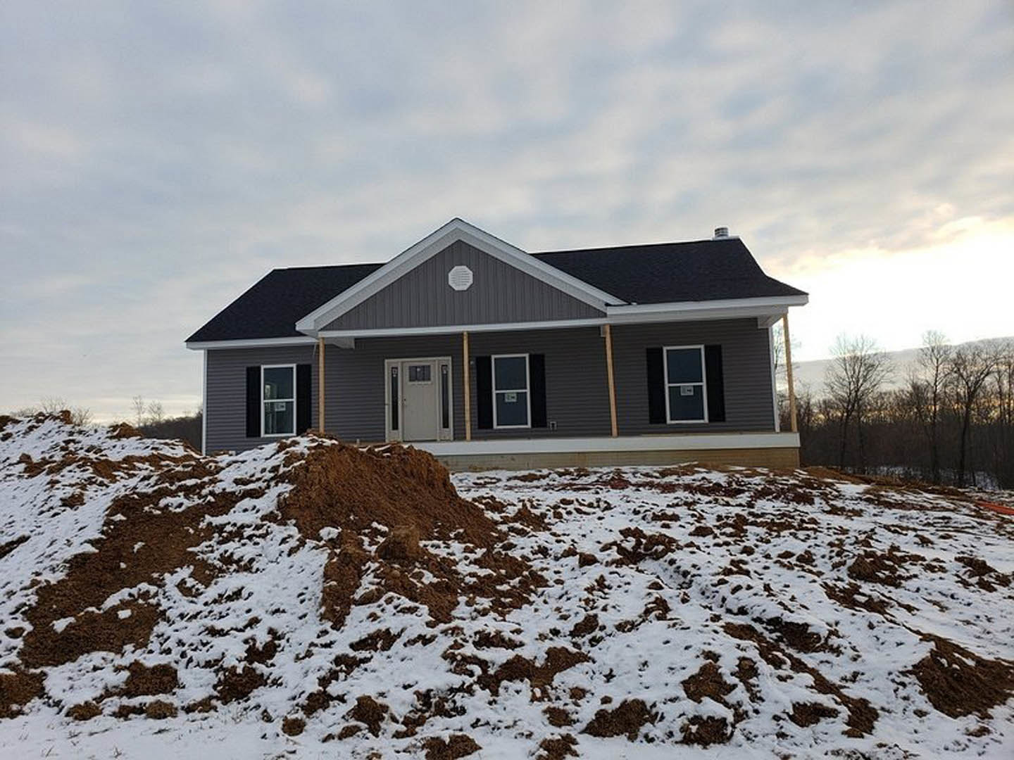 Two-story cottage with white door and window, surrounded by snow-covered ground and leafless trees, cloudy winter sky overhead.