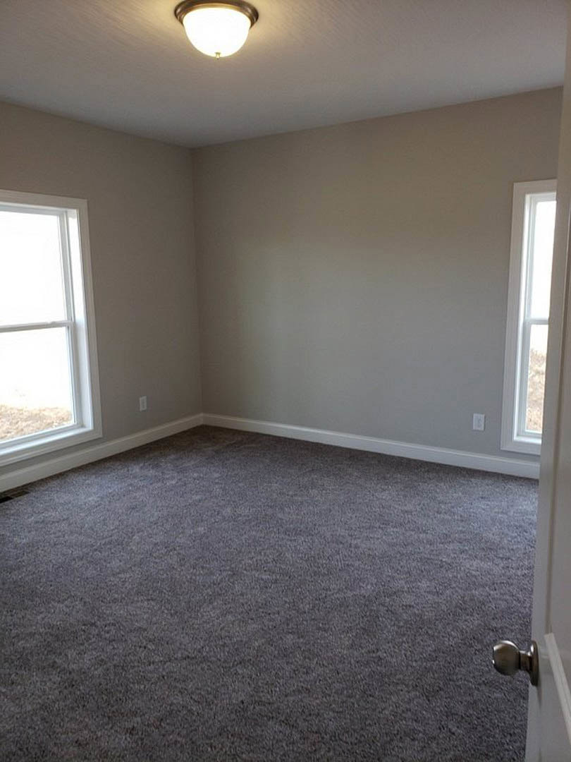Grey carpeted room with white plaster walls, ceiling-mounted light fixture, and large window letting in natural light