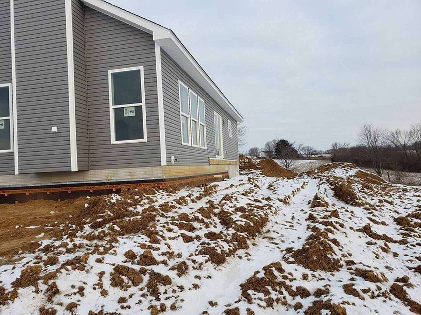 Grey house with white trim, snow covering the ground, dirt pile and brown debris near the foundation, window displaying a sign, winter trees and cloudy sky in the background